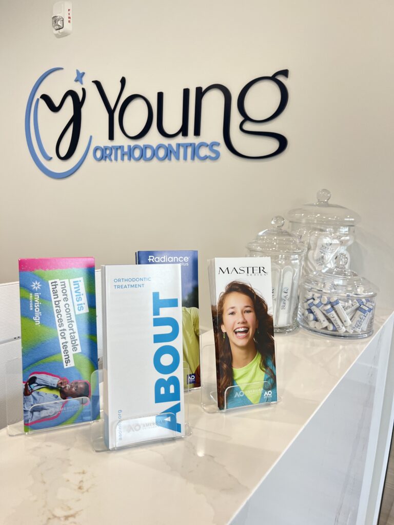 A reception counter at Young Orthodontics featuring three informational brochures and glass jars filled with dental hygiene supplies. The brochures are displayed upright in clear plastic holders against a wall displaying the Young Orthodontics logo.